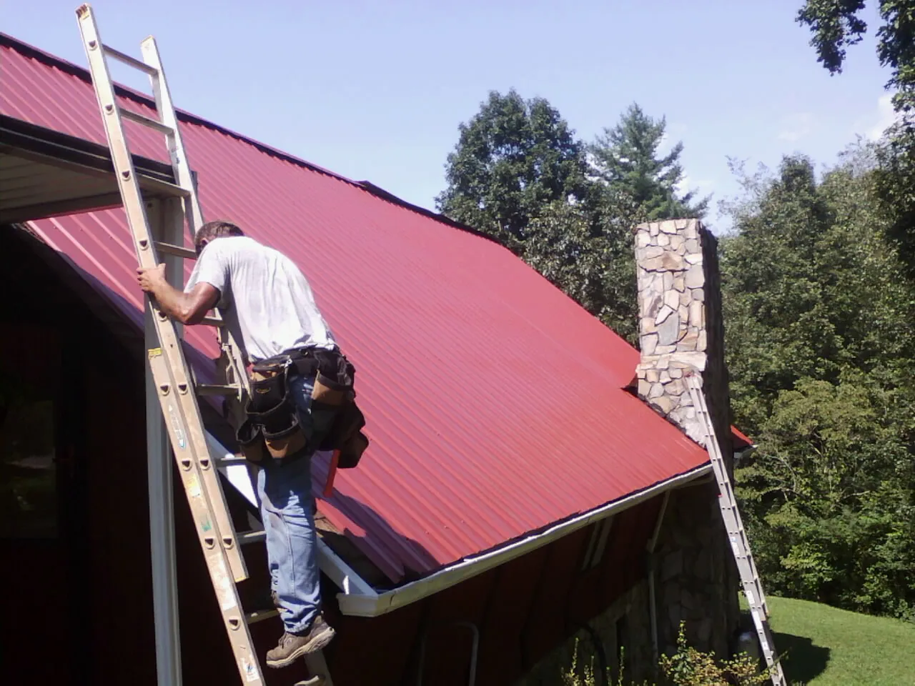 Roofer on a ladder beside a red metal roof and stone chimney — Soffit & Fascia Repair by Roofing Brothers in Indian Springs